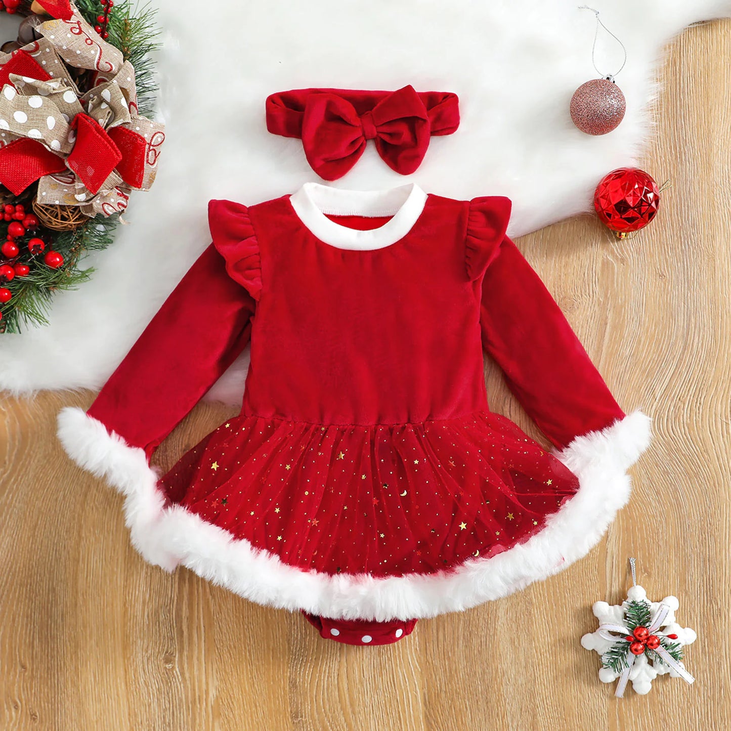 Top-down image of red romper and bow arranged with pine branches and star ornament on wooden background.